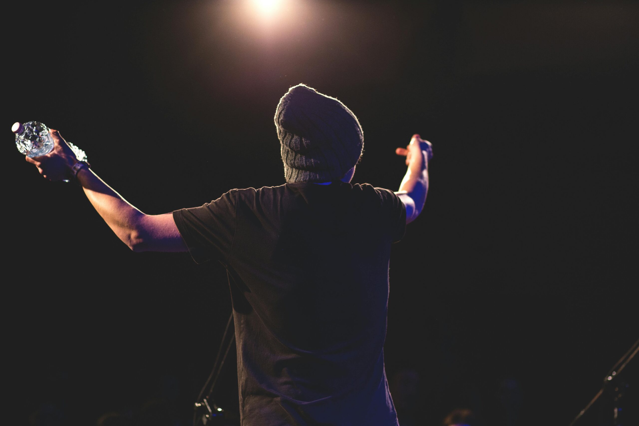 Back view of a musician performing energetically on stage under bright lights with an audience.