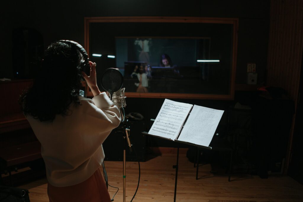 A female singer records vocals in a professional music studio with a microphone and sheet music.