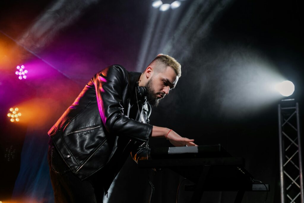 DJ in a leather jacket performing on stage with vibrant colored lights at a concert.