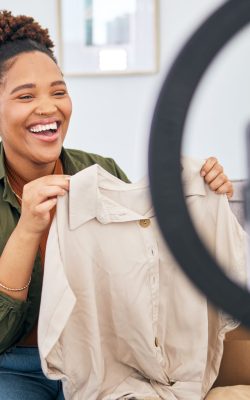 Fashion, live streaming and a black woman content creator unboxing a clothes outfit in her home. Social media, brand deal and a happy female influencer recording a broadcast for subscription service.