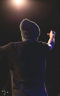Back view of a musician performing energetically on stage under bright lights with an audience.